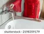 Small photo of A woman in red silk nightgown turns on the faucet at the bathtub. Women prepare for taking a bath