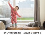 A toddler wearing pink pyjamas, long socks and slippers exercises on top of a wicker stool and sticks her tongue out while her British Short Hair cat sits next to her in a house in Edinburgh, Scotland