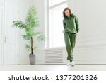 Full length shot of active pleased young woman with slim body, keeps hands in pockets of tracksuit, focused down, ready for doing cardio exercises, has determined look, pilates exercising indoor