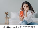Small photo of Stressed caucasian girl is reading morning news looking at smartphone display. Young woman is chatting on phone or browsing internet at home in her bedroom. Lady is doom scrolling.