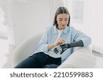 Small photo of Beautiful young girl with disability setting her sensory bionic prosthetic arm, sitting in armchair. Modern woman using artificial robotic hand after limb loss.