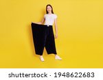 Small photo of Slim attractive girl with dark hair wearing white caucasian t shirt and too big black pants, female lost weight and being proud of it, looks smiling at camera, isolated over yellow background.