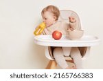 Small photo of Image of thirsty toddler baby girl dresses in beige jumper sitting in high chair and drinking water from yellow bottle, looking aside, studying area around her, posing isolated over white background