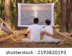 Small photo of Outdoor shot of lovely couple sitting in the forest with overhead projector, watching movie or photos from vacation, hugging each other and enjoying time together.