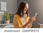 Portrait of adorable positive woman with brown hair wearing yellow shirt and eyeglasses posing in office, holding mobile phone in hands, typing on smart phone.
