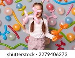 Indoor shot of little girl with braids wearing casual clothing posing isolated over gray background with balloons, holding air ballons animal figures, playing on party.