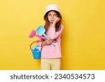 Small photo of Bored sad little girl wearing casual clothing and panama holding sandbox toys and bucket isolated over yellow background feeling disinterest at playground posing with rake and scoop.