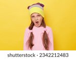 Small photo of Deleterious little girl with ponytails wearing pink shirt and beanie hat standing isolated over yellow background screaming expressing negative emotions.