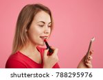 Sideways portrait of female lady paints lips with red lipstick, uses smart phone as mirror, prepares for date with handsome man, applies cosmetic, isolated over pink background. Beauty concept