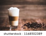 Caffe latte layered with milk in a high drinking glass. There are roasted coffee beans spread out on the wooden table next to the glass, and the background is also wooden.