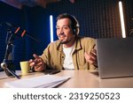 Young stylish man in mustard shirt with headphones gesturing at microphone and sharing story with audience while sitting at desk in studio with neon lighting and recording podcast