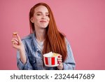 cute red long foxy haired ginger woman holding packaging cardboard box bucket with french fries and eating chicken crunchy nuggets in pink studio
