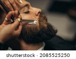 Small photo of Young bearded man getting shaved with straight edge razor by hairdresser at barbershop