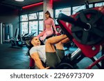 Two muscular gym mates are doing leg exercises on the leg press machines in a gym in different positions. Selective focus on a sportswoman. Sporty couple working out in a gym on the machines.