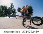 Small photo of A shirtless tattooed mature man is performing tricks and stunts in a skate park with his bmx. A mature urban freestyle bike rider is practicing tricks and stunts on his bike in a skate park.
