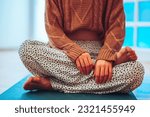 Small photo of Cropped shot of a caucasian girl in a dotty yoga pants sitting on a mat in a lotus pose with her hands crossed forward. Wearing a knitted sweater and long legged pants while enjoying her morning yoga.