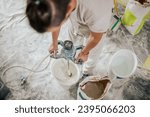 Small photo of A builder is mixing plaster in a bucket with manual cement mixer while standing in a messy house in a renovation process. A constructor is making materials for plastering and skim coating walls.