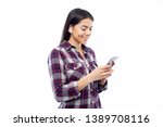 Portrait of young female student, holding mobile phone in both hands, wearing wireless earphones, looking at the screen and smiling, wearing casual plaid shirt, isolated on white background