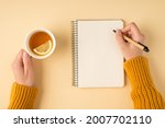 First person top view photo of female hands in yellow pullover writing in spiral planner and holding white cup of tea with lemon slice on isolated pastel orange background with copyspace