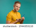 Handsome young man making notes in planner, guy holding pen, reflects on plans. Guy in yellow wear makes records in diary on blue studio background.