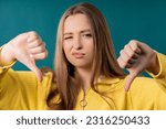 Small photo of Portrait of unhappy european woman condemns with sign of dislike. Young millennial lady expressing discontent with showing thumbs-down gesture on blue studio background.