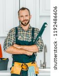 Small photo of handsome plumber holding monkey wrench and looking at camera in kitchen