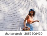 cheerful young african american woman in sunglasses and headscarf holding longboard near brick wall