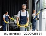 happy man with floor scrubber machine looking at camera near multiethnic women cleaning office