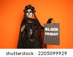 happy woman in mexican day of dead makeup and costume holding black friday shopping bag isolated on orange