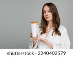 young woman with shiny hair holding bottle with shampoo isolated on grey