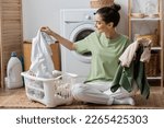 Happy young woman sorting clothes near washing machine in laundry room