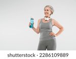 carefree senior woman with wireless headphones holding sports bottle and standing with hand on hip isolated on grey
