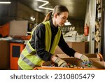 Young female worker of garbage sorting center wearing protective clothing and gloves while working with plastic caps in carton boxes, garbage sorting and recycling concept