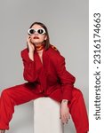 Small photo of generation z, fashion model with brunette short hair and nose piercing posing in sunglasses and red suit while sitting on concrete cube on grey background, lady in red, young woman in red outfit