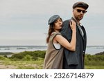 Small photo of Portrait of brunette woman in newsboy cap and vest embracing bearded boyfriend in sunglasses and jacket while standing with blurred landscape at background, trendy twosome in rustic setting