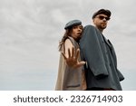 Small photo of Low angle view of fashionable brunette woman in newsboy cap and vest looking at camera near boyfriend in jacket and sunglasses with cloudy sky at background, trendy twosome in rustic setting