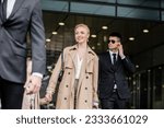 Small photo of professional bodyguards protecting smiling blonde woman and preteen kid, successful mother and daughter in trench coats standing near hotel, safety and protection, private security, guards