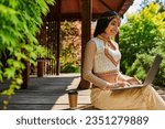 Small photo of summer park, happy ethnic style woman typing on laptop near coffee to go in wooden alcove