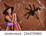girl in witch hat and Halloween costume grimacing near fake spider and cobwebs on brown background