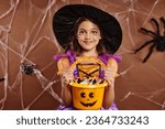 happy girl in witch hat with spiderweb makeup holding bucket of sweets on brown with spider net