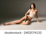 beautiful asian model in corset and beige fishnet tights sitting while posing on grey background