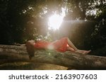 a young and beautiful woman in a red summer dress with white flecks lying on a fallen dry tree trunk in the middle of the forest