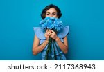 Small photo of Portrait of a gorgeous woman, who is looking in the camera and hiding a part of her face over the bunch of blue flowers. Mothers day. Women's holidays. Springtime. Women rights. The 8th of March