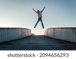 Young caucasian man in jeans and hoodie jumping with spread out arms on concrete bridge. Mid air parkour pose in city environment and clear sky