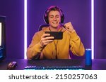 Playing video games on smartphone. Young caucasian handsome man sitting on chair holding cellphone in his hand. Exited streamer wearing headset celebrating victory in online mobile game in neon room