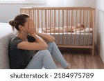 Side view of mother covering face crying from exhaustion sitting on floor next to baby crib while child napping, can't handle difficulties of maternity, having postnatal depression symptoms
