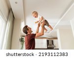 Young dad throwing excited laughing baby boy up in air and catching, amusing kid and having fun together after returning home from work while mother cooking dinner. Active play at home