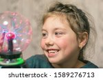 Portrait of a cheerful little girl playing with a plasma ball. The child smiles and laughs while looking at the lightning in a decorative round lamp. Selective focus.