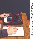 Small photo of close up male songwriter hands writing a hit song on white paper while playing music keyboard synthesizer. songwriting concept