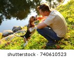 Small photo of Man sit on squat on haunches at green bank of river, play with young beagle dog. Owner tease doggy with wooden stick, pet stand on hind legs and gnaw twig